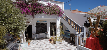 Un encantador patio con paredes blancas, vibrantes buganvillas, suelo de baldosas, escalera exterior, acogedores asientos, plantas en macetas, menú de pizarra y una brillante luz solar bajo un cielo azul despejado.