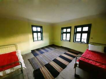 L'image représente une chambre avec deux lits, des murs jaunes et trois fenêtres, avec un tapis au sol et un plafond blanc.