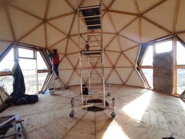 L'image représente un dôme géodésique en construction, avec une échelle, un plancher en bois et des fenêtres, avec une personne debout sur une échelle à l'arrière-plan, entourée de lumière naturelle.