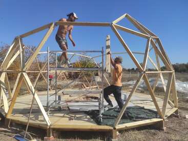 Deux hommes construisent un dôme géodésique par une journée ensoleillée, l'un se tenant sur un échafaudage et l'autre tenant une pièce de charpente en bois.