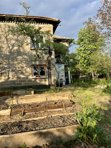 Une vieille maison au toit de tuiles se dresse à côté d'un jardin agrémenté de plates-bandes surélevées, d'arbres et d'une végétation luxuriante, baignée par la douce lumière du soleil sous un ciel nuageux.