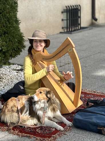 Une femme coiffée d'un chapeau joue de la harpe en bois, assise sur un tapis à motifs à l'extérieur. Un chien au pelage duveteux est couché à ses côtés ; tous deux profitent du soleil.