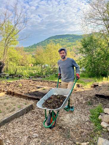 Un homme pousse une brouette remplie de déchets de jardin dans un jardin verdoyant. Derrière lui, on aperçoit des plates-bandes surélevées et une montagne pittoresque.