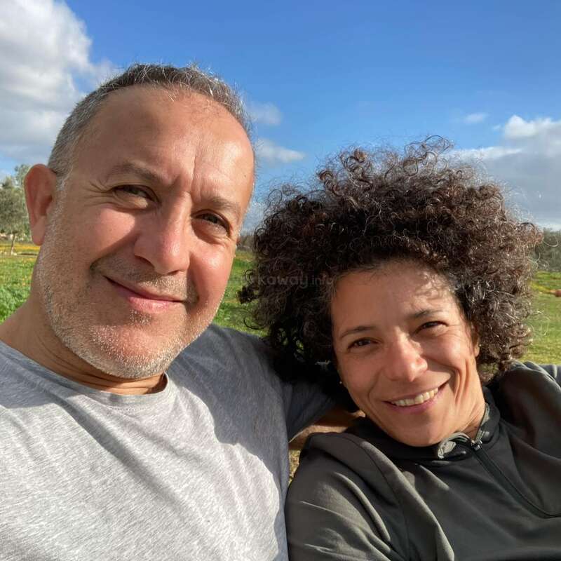 The image shows a man and woman posing for a selfie in a grassy field with trees and a blue sky with white clouds in the background.