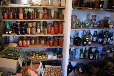 Una despensa rústica repleta de tarros de verduras y frutas en conserva, botellas de vino, patatas, manzanas, hierbas aromáticas y otros productos caseros colocados en estantes de madera, que parece estar bien surtida.