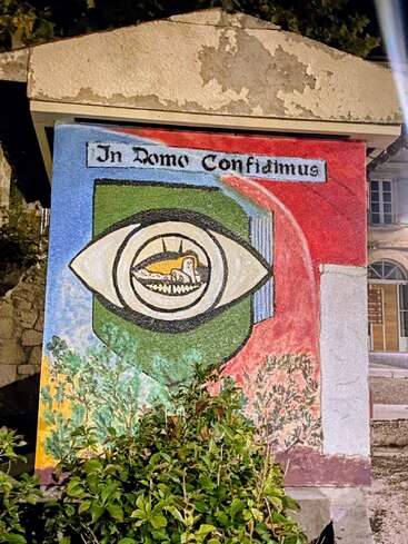 Esta imagen muestra un colorido mural en el que aparecen un ojo, un cordero y la frase en latín «In Domo Confidimus», rodeado de vegetación y bajo la luz nocturna.