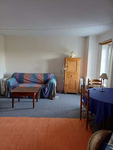 Ein gemütliches Wohnzimmer mit einem bunt gestreiften Sofabezug, einem Couchtisch aus Holz, einem Schrank, einer Stehlampe, einem runden Tisch mit blauem Tischtuch, Stühlen und einem großen Fenster.