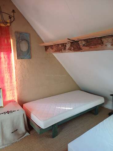 Ein gemütliches Dachzimmer mit einem Einzelbett mit weißer Matratze, rustikalen Holzbalken, einem kleinen Tisch, einem roten Vorhang, abstrakter Wandkunst und warmer Beleuchtung.