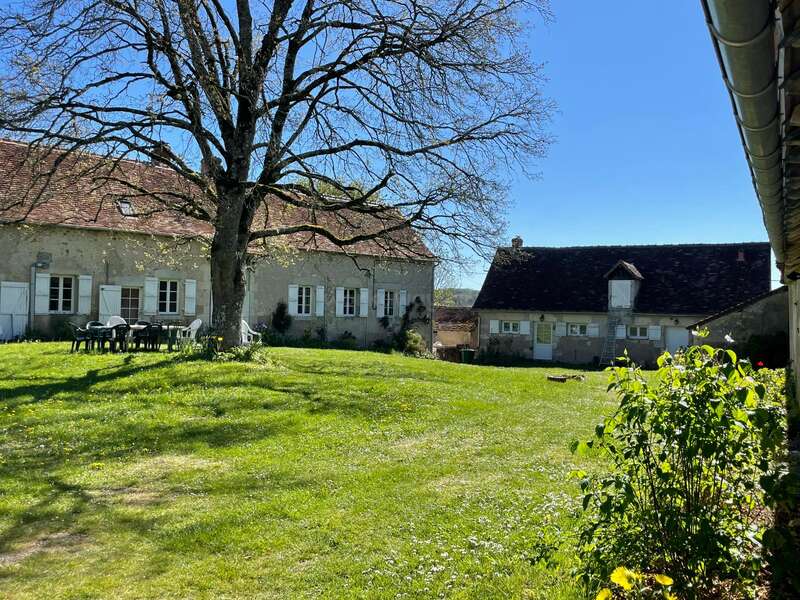 Eine friedliche ländliche Szene mit zwei Steinhäusern, einem großen Baum, einem grünen Rasen, einem Tisch und Stühlen im Freien, unter einem strahlend blauen Himmel an einem sonnigen Tag.