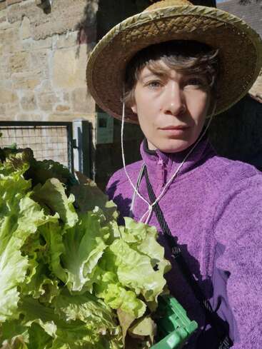 Eine Person, die einen Strohhut und einen lila Pullover trägt, hält einen frisch geernteten Salat in der Hand und steht an einem hellen, sonnigen Tag im Freien an einer Steinmauer.