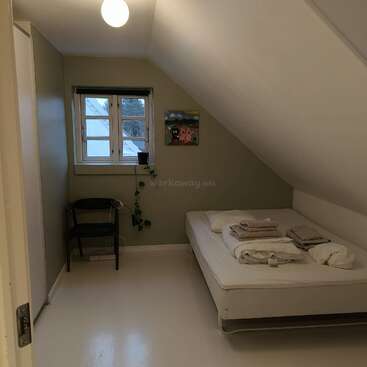 A minimalist bedroom with slanted ceiling, single bed, fresh linens, small window, chair, hanging plant, wall art, neutral colors, soft lighting, and clean, serene atmosphere.