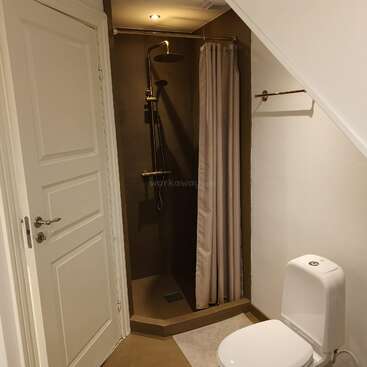 This image shows a compact bathroom with a white toilet, a small shower with a curtain, a towel bar, and a white paneled door under a sloped ceiling.