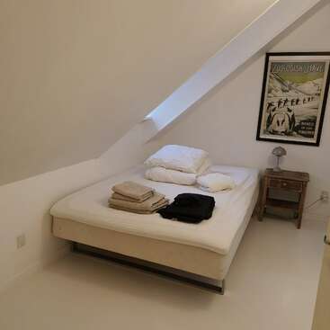 A minimalist bedroom with slanted white walls, a bed with folded linens and clothes, a wooden nightstand, a lamp, and a penguin-themed poster. Cozy atmosphere.