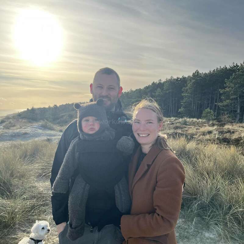 A smiling family stands together outdoors in a grassy, sandy landscape. The father holds a bundled-up baby, while a small white dog sits nearby under the bright sun.