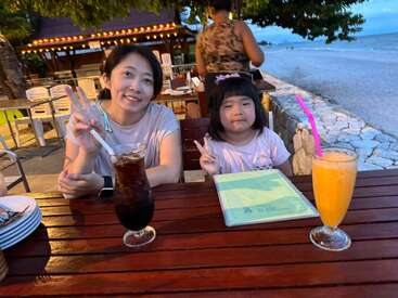A woman and a young girl sit at a beachside restaurant, smiling and making peace signs, with refreshing drinks, menus, and the ocean in the background.