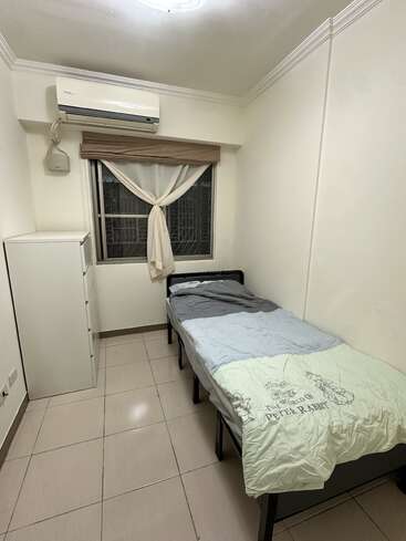 A small, tidy bedroom with cream walls, a single bed, "The World of Peter Rabbit" bedding, a white dresser, window with tied curtains, and air conditioner.