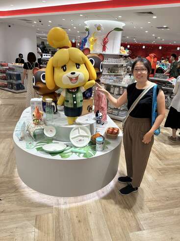 A woman stands smiling next to large Animal Crossing character figures in a brightly lit store filled with merchandise and colorful decorations, with other shoppers browsing.