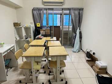 This image shows a small classroom with six wooden desks and chairs, some cluttered shelves, boxes, and a window with closed curtains. The room appears tidy.