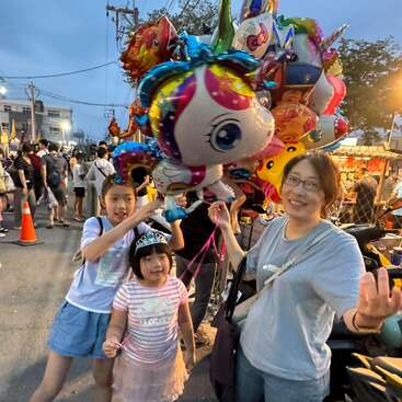 Una mujer y dos niños están de pie en un mercado nocturno al aire libre, sonriendo y sosteniendo globos de unicornios de colores. Multitudes, luces y vendedores ambulantes llenan el animado fondo.