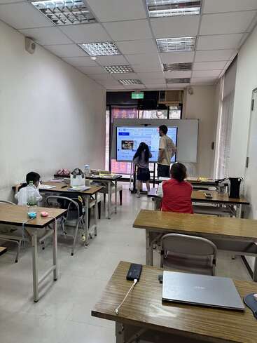 Uma sala de aula com vários alunos sentados nas carteiras, enquanto um aluno e um professor interagem com uma grande tela sensível ao toque na frente da sala.