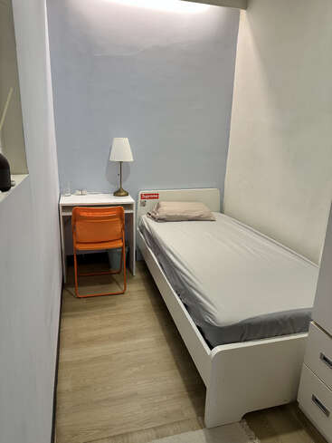 Este é um quarto pequeno e minimalista, com uma cama de solteiro, roupa de cama cinza claro, paredes brancas, uma cadeira laranja, um abajur e piso de madeira.