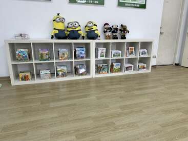 A área de leitura infantil conta com estantes brancas com cubículos repletas de livros ilustrados coloridos, além de peluches dos Minions e ursinhos de pelúcia dispostos de forma alegre em cima delas.