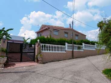 The image depicts a two-story house with a brown brick exterior, a white fence, and a black gate, set against a blue sky with white clouds.