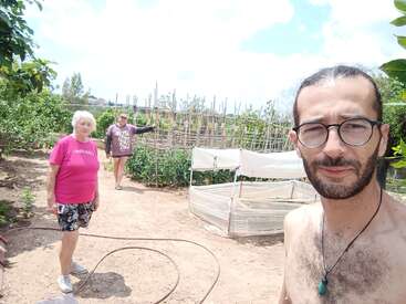 The image depicts three individuals in a garden setting, with two standing on a dirt path and one partially visible in the foreground. The scene is set against a backdrop of lush greenery.