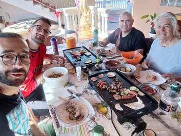 The image shows four people sitting at a table with food and drinks, smiling for a selfie. They are enjoying a meal together.