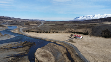 Uma casa branca solitária com telhado vermelho fica às margens de um rio sinuoso, cercada por campos áridos, montanhas distantes cobertas de neve e um céu azul límpido.