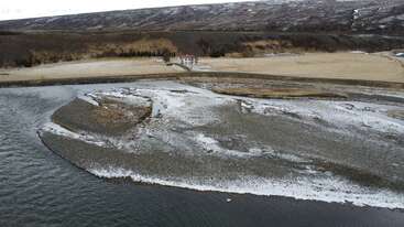 Um rio serpenteia por uma paisagem rochosa parcialmente coberta de neve. Ao fundo, uma casa fica isolada em um campo gramado, perto de uma encosta íngreme e árida.