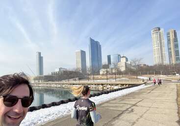 A man in sunglasses smiles at the camera while people jog on a pathway beside water, with snow on the ground and tall city buildings in the background.