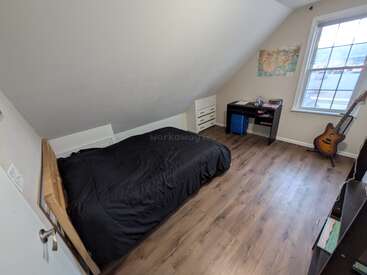 This cozy bedroom features a single bed with a black comforter, a small desk, dresser, world map, wooden floors, and a guitar near a large window.