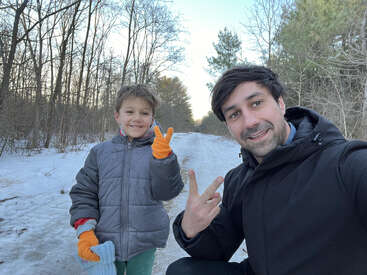 Un homme et un enfant profitent d'une promenade hivernale sur un sentier enneigé dans les bois ; tous deux sourient et font le signe de la paix avec leurs mains gantées.