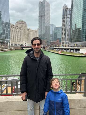 A man and a smiling boy stand in front of a green river, surrounded by tall city skyscrapers on a cloudy day, enjoying their time outdoors.