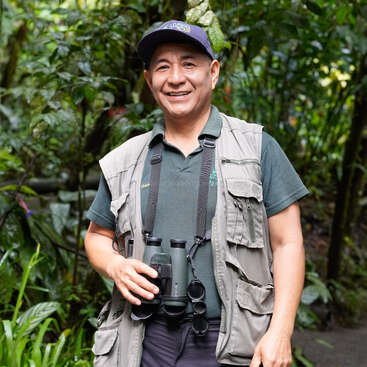 A smiling man stands in a lush forest, wearing a cap, green polo, and khaki vest. He holds binoculars, appearing ready for birdwatching or nature exploration.