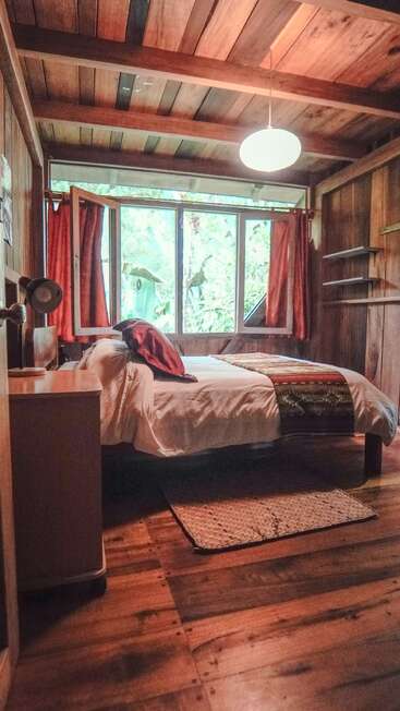 This cozy wooden bedroom features a double bed with white linens, red accents, large windows with red curtains, wooden ceiling, small desk, and natural light. Peaceful atmosphere.