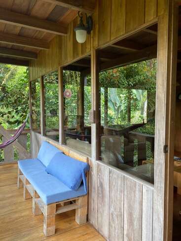 A cozy wooden porch with large windows, a blue-cushioned bench, and a hammock. Surrounded by lush greenery, it offers a peaceful, inviting outdoor retreat.