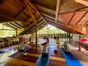 A group of people practice yoga or meditate in a spacious, rustic wooden loft with natural light. The setting feels peaceful, surrounded by lush greenery outside.