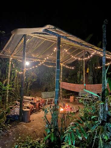 A cozy outdoor shelter with a bamboo roof, string lights, benches, firewood, and a campfire. Surrounded by lush greenery, creating a warm, inviting nighttime atmosphere.