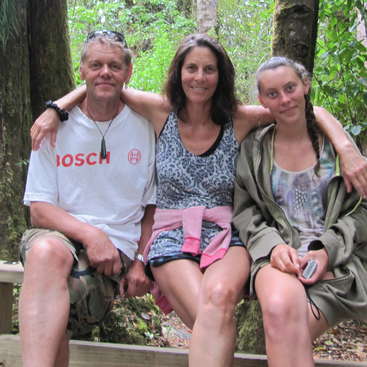 The image shows a man, woman, and girl sitting on a wooden bench in a wooded area, posing for a photo with their arms around each other.