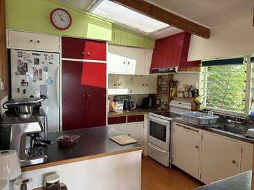 This kitchen features red and white cabinets, a white stove, refrigerator, coffee machine, counter appliances, cutlery, a clock, wood accents, dishes, and ample sunlight.