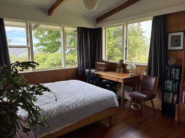 This cozy bedroom features large windows with tree views, a wooden bed with white bedding, a desk, bookshelves, indoor plant, and wooden beams on the ceiling.