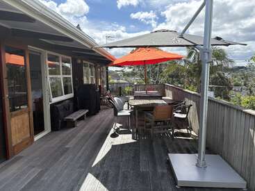 A sunny outdoor wooden deck attached to a house, featuring tables, chairs, two large umbrellas for shade, lush greenery, and a distant scenic neighborhood view.