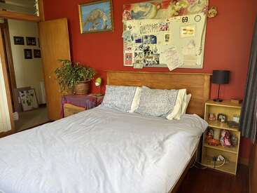 This cozy bedroom features a wooden bed, soft pillows, red accent wall, plants, bedside shelves, personal photos, art, and natural light from an open door.