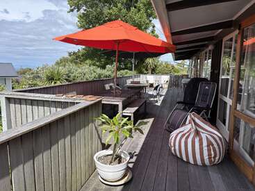 A wooden deck features a large red umbrella, outdoor seating, a striped beanbag, potted plants, and is surrounded by greenery. Glass doors open into the house.