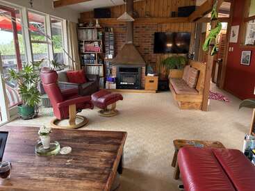 This cozy living room features a rustic fireplace, wood-paneled walls, vintage furniture, houseplants, a large window, bookshelves, and a television for comfortable, inviting relaxation.
