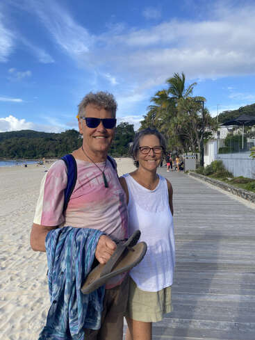 A smiling couple stands on a boardwalk by the beach, dressed casually. Palm trees, blue sky, and sand create a relaxed, tropical, and happy atmosphere.