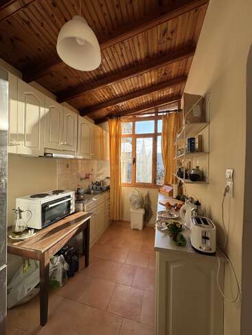 Une cuisine étroite et confortable avec des placards couleur crème, un plafond en bois et un sol carrelé. La lumière du soleil pénètre à flots par les grandes fenêtres aux rideaux jaunes. Des appareils électroménagers et de la vaisselle sont éparpillés un peu partout.