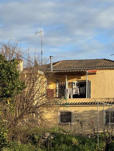 Une maison jaune avec un toit en tuiles et un balcon où sèche du linge. Des arbres feuillus et de la verdure entourent la maison, sous un ciel partiellement nuageux.